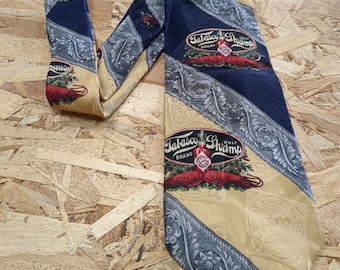 May include: A yellow and blue patterned silk tie with a Tabasco Shrimp logo. The logo features a red bottle of Tabasco sauce and a shrimp. The tie has a silver and blue floral pattern.