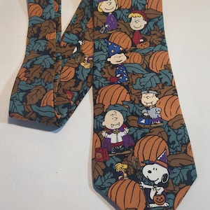 May include: A necktie featuring a repeating pattern of Peanuts characters in Halloween costumes, including Snoopy, Charlie Brown, and Lucy, surrounded by orange pumpkins and dark teal autumn leaves on a dark background.