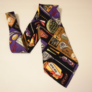 Puede incluir: Una corbata de seda morada y negra con un patr&oacute;n de tem&aacute;tica de golf. La corbata presenta palos de golf, pelotas de golf y las palabras "Top-Flite" y "The Air."