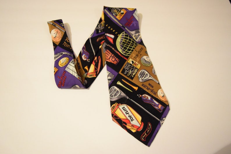 Puede incluir: Una corbata de seda morada y negra con un patr&oacute;n de tem&aacute;tica de golf. La corbata presenta palos de golf, pelotas de golf y las palabras "Top-Flite" y "The Air."