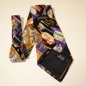 Puede incluir: Una corbata de seda morada y dorada con tem&aacute;tica de golf. La corbata presenta im&aacute;genes de palos de golf, pelotas de golf y el texto "Top-Flite" repetido en toda la superficie. La corbata est&aacute; hecha en Corea.