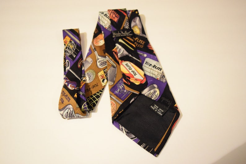 Puede incluir: Una corbata de seda morada y dorada con tem&aacute;tica de golf. La corbata presenta im&aacute;genes de palos de golf, pelotas de golf y el texto "Top-Flite" repetido en toda la superficie. La corbata est&aacute; hecha en Corea.