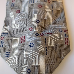 May include: A gray necktie with a repeating pattern of white, gray, and brown lines, resembling a circuit board. Small clusters of red and blue dots and flower-like shapes are interspersed throughout the design. The tie is made of a textured fabric.
