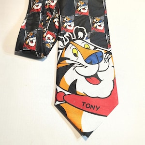 May include: A black necktie featuring a repeating pattern of a cartoon tiger in a red and white square. The tie's tip showcases a large tiger face with the name "TONY" on a red collar.