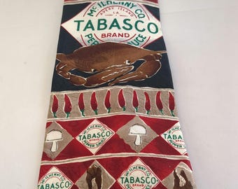 May include: A dark blue silk tie with a repeating pattern of red and white diamonds. The tie features a red and white logo for McIlhenny Company's Tabasco brand pepper sauce, a crab, mushrooms, and red peppers. The text on the logo reads "McIlhenny Company Avery Island, LA Tabasco Brand Pepper Sauce."