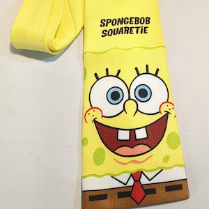 May include: Yellow necktie featuring a smiling cartoon character face with blue eyes, a red mouth, and white teeth. The tie has the words "SPONGEBOB SQUAREPANTS" printed on it. The tie is yellow with a repeating pattern.