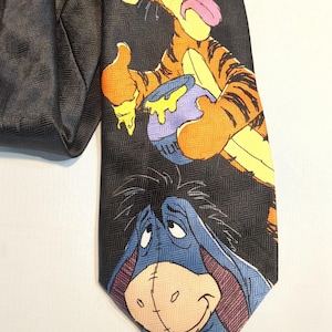 Puede incluir: Corbata negra con Tigger y Eeyore de Winnie the Pooh. Tigger es naranja y negro, sosteniendo un tarro de miel. Eeyore es azul con una expresión triste. La corbata tiene un patrón de espiga negro.