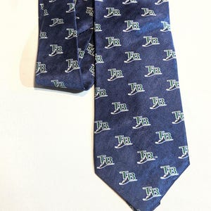 May include: Navy blue necktie with a repeating pattern of the letters "TB" in white with green accents. The tie is made of a smooth, possibly silk, material and is displayed on a white surface.
