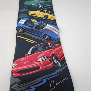 Op de afbeelding: Een zwarte stropdas met een herhaald patroon van kleurrijke sportwagens. De auto's zijn rood, geel, groen, wit en blauw. De stropdas heeft de handtekening "Simon" in witte letters.