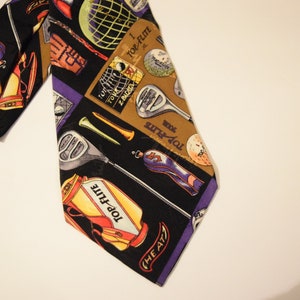 Puede incluir: Una corbata de seda negra con un patr&oacute;n colorido de palos de golf, pelotas de golf y bolsas de golf. La corbata tiene el texto "Top-Flight" y "The A77".