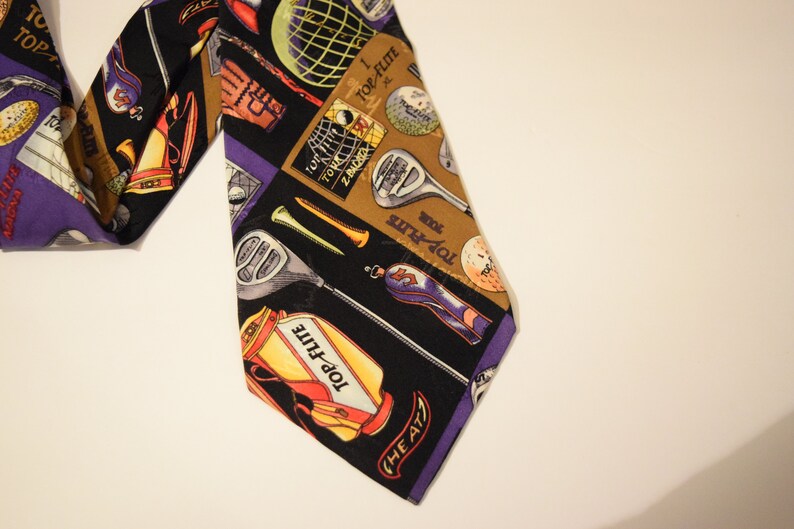 Puede incluir: Una corbata de seda negra con un patr&oacute;n colorido de palos de golf, pelotas de golf y bolsas de golf. La corbata tiene el texto "Top-Flight" y "The A77".