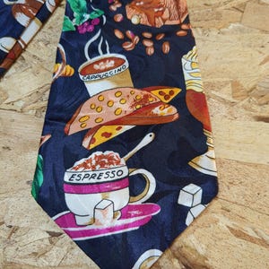 May include: A dark blue silk tie with a colorful pattern of coffee cups, coffee beans, pastries, and sugar cubes. The tie features the words "Cappuccino" and "Espresso" on the cups.