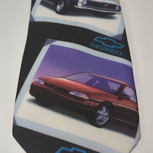 May include: Black necktie featuring images of classic Chevrolet cars in white-bordered frames. The tie includes the Chevrolet logo in blue. The cars are in various colors, including red, blue, and green.