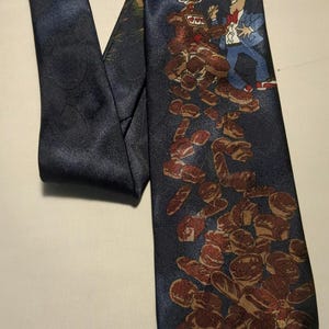 Op de afbeelding: Een blauwe zijden stropdas met een cartoonillustratie van een man die op een stapel bruine gebakjes staat. De stropdas heeft een goud- en bruin patroon.