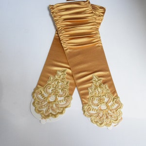 May include: A pair of long, gold satin gloves with delicate lace detailing at the wrists. The lace is a lighter shade of gold and features a floral design.