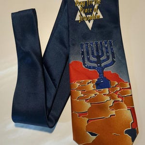 Pode incluir: Uma gravata azul marinho com um design dourado e vermelho que apresenta um candelabro e as palavras "Pray for the Peace of Jerusalem" dentro de uma estrela de David branca.
