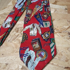 Puede incluir: Corbata de seda roja con un patrón de varios escudos de armas universitarios, libros y otros símbolos académicos. La corbata está hecha de una tela suave y lisa y tiene un diseño clásico.