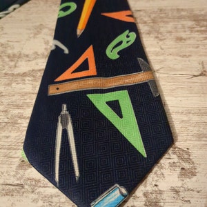 May include: Navy blue necktie with a repeating pattern of school supplies. The tie features orange triangles, green curves, a yellow pencil, and a ruler. A compass and a blue eraser are also visible.
