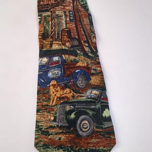 May include: A patterned necktie with a rural scene featuring a cabin, an American flag, vintage trucks, and dogs. The tie's design incorporates shades of brown, green, and blue, creating a detailed and thematic accessory.