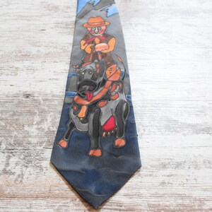 May include: A blue and gray silk tie with a cartoon illustration of a person riding a bull. The person is wearing a brown hat and orange shirt. The bull is black and gray with a red and orange saddle.