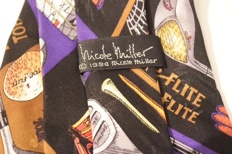 Puede incluir: Una corbata de seda negra con tem&aacute;tica de golf. La corbata presenta una variedad de palos de golf, pelotas de golf y las palabras "Top Flite" y "Elite". La corbata tambi&eacute;n tiene una etiqueta que dice "Nicole Miller" y "&copy; 1994 Nicole Miller".