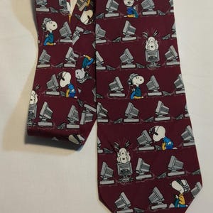 May include: A burgundy tie featuring a repeating pattern of Snoopy characters and computer monitors. The cartoon characters are in various poses, some wearing headphones. The tie is made of a smooth fabric and is suitable for a business or casual setting.
