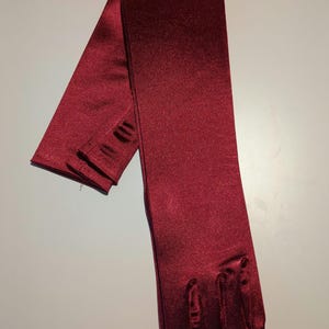Puede incluir: Un par de guantes largos de satén burdeos. Los guantes son sin dedos y tienen una textura suave y sedosa.