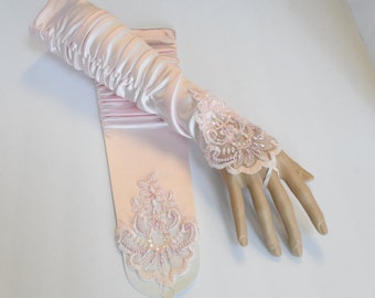 May include: A pair of pale pink satin opera-length gloves featuring ruched detailing along the forearm and an intricate light pink lace appliqué on the back of the hand and wrist, embellished with small beads and iridescent sequins.