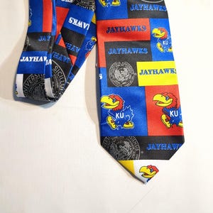 May include: A blue, red, and yellow patterned necktie featuring the text "JAYHAWKS" and the University of Kansas Jayhawk mascot. The tie has a repeating pattern of squares with the Jayhawk logo and university seals.