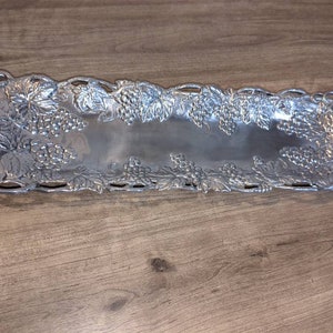 Könnte beinhalten: Ein silbernes Serviertablett mit einem Traubenranken-Design. Das Tablett ist rechteckig und hat einen erhöhten Rand.