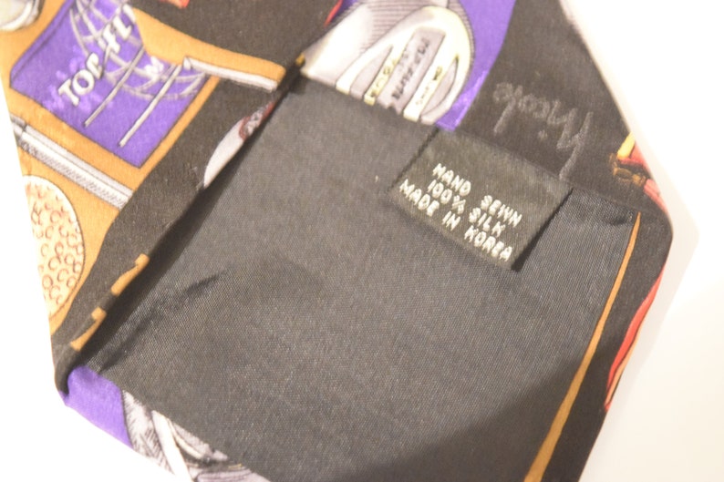 Puede incluir: Una corbata de seda negra con un patr&oacute;n colorido de im&aacute;genes relacionadas con el golf, incluyendo pelotas de golf, palos de golf y las palabras "Top-Flight". La corbata tiene una etiqueta que dice "Hand Sewn 100% Silk Made in Korea."