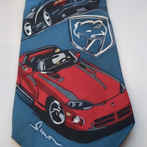 Puede incluir: Una corbata azul con ilustraciones de coches deportivos rojos y negros. La corbata tiene un patrón repetido de gráficos de coches, un emblema plateado y una firma. Los coches son de estilo carreras.