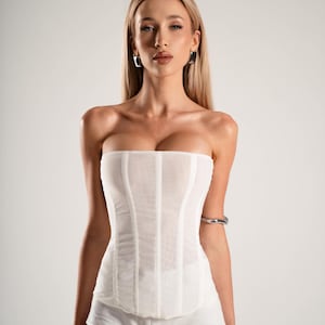 May include: A white strapless corset top with vertical paneling and matching white shorts. The model is wearing silver hoop earrings and a silver arm cuff. The background is a solid white.