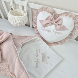 Può includere: Un cuscino bianco a forma di cuore con fiocco rosa e bordi in pizzo, ricamato con "Everly Grace". Sono visibili anche una coperta per bambini abbinata e un cestino all'uncinetto, che suggeriscono un tema per la cameretta.