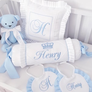 Puede incluir: Un juego de ropa de cama para bebé en una cuna, con un osito de peluche azul, una almohada con una "H" bordada, un cojín cilíndrico con "Henry" y una corona, y dos baberos. El conjunto es blanco con detalles azules.