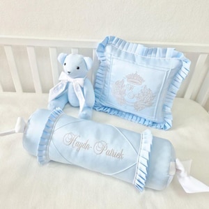 May include: A baby blue nursery set featuring a teddy bear, a decorative pillow, and a bolster pillow. The pillow and bolster are embroidered with a crown and the name "Haydn-Patriek". The set is trimmed with white ruffles and bows.
