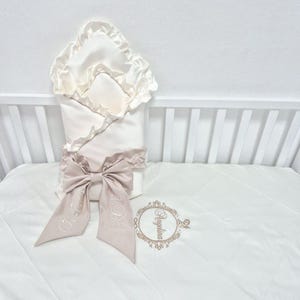 May include: A cream-coloured baby swaddle blanket with a matching bow and decorative trim. The bow features embroidered initials. The blanket is placed on a white cot with a matching white cot sheet. The sheet has the name "Angelina" embroidered on it.