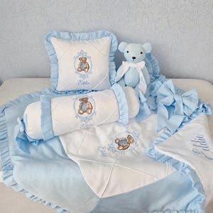 Può includere: Un set di biancheria da letto per bambini in azzurro e bianco, con un motivo a orsetto. Il set include un cuscino, un cuscino a rullo, una coperta e un peluche a forma di orsetto. Gli articoli hanno bordi a balze e il nome "Eddie" è ricamato.