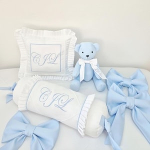 May include: A collection of baby bedding and accessories in soft blue and white. Includes a plush teddy bear, decorative bows, a square pillow, and a bolster pillow, all with embroidered initials. The items are arranged on a white surface.