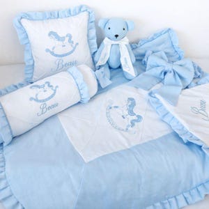 Puede incluir: Un juego de ropa de cama para bebé en azul claro y blanco, con un diseño de caballito balancín. El juego incluye una almohada, un cojín cilíndrico, una manta y un osito de peluche, todos bordados con el nombre "Beau". La manta tiene un borde con volantes.