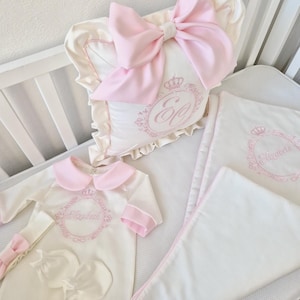 May include: A baby bedding set in a cot, featuring a white and pink colour scheme. The set includes a pillow with a large pink bow, a blanket, and a onesie, all embroidered with a crown and a name. The overall theme is elegant and delicate.
