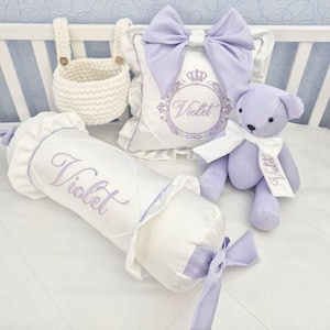 May include: A nursery set featuring a white and lavender color scheme. Includes a white crochet basket, a bolster pillow, a decorative pillow with a bow, and a teddy bear. The name "Violet" is embroidered on the pillow, bolster, and bear.