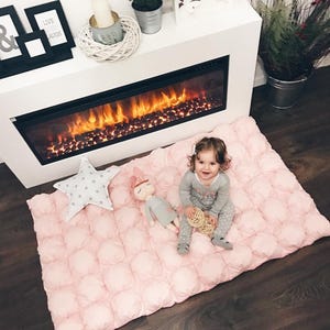 May include: A pink, quilted baby play mat with a star-shaped pillow and a doll. A child in gray pajamas sits on the mat near a fireplace. The fireplace has a black frame and a glowing fire effect.