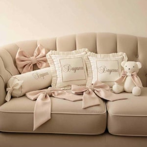 Puede incluir: Un sofá beige exhibe cojines decorativos, lazos y un oso de peluche. Los cojines cuadrados tienen bordes con volantes y el nombre "Dayana" bordado. Una almohada cilíndrica y grandes lazos en tonos rosa suave complementan el conjunto.