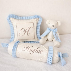 May include: A cream-colored nursery set featuring a square pillow with the letter "H", a bolster pillow with the name "Hughie", and a teddy bear. All items have light blue accents and embroidery.