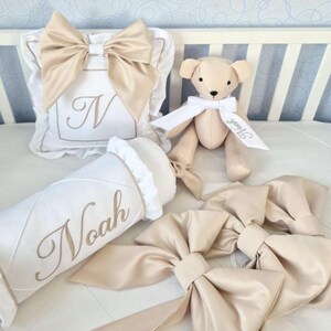 May include: A nursery set featuring a white pillow with a large beige bow and the letter "N", a beige teddy bear with a white bow, a white bolster pillow embroidered with "Noah", and several beige bows. The items are arranged on a white surface.