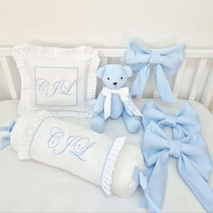 Puede incluir: Un juego de cuna para bebé con un osito de peluche azul claro, dos lazos grandes, una almohada y un cojín cilíndrico. El juego es blanco con detalles en azul claro e iniciales bordadas.