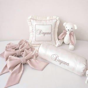 May include: A collection of baby items in soft pink and white. Includes a white teddy bear with a pink bow, a square pillow and a bolster pillow, both embroidered with the name "Dayana", and two large pink bows.