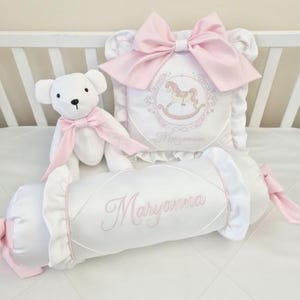 Puede incluir: Un oso de peluche blanco con un lazo rosa está al lado de una almohada blanca con un lazo rosa y un diseño de caballito balancín. También hay un cojín cilíndrico blanco con el nombre "Maryanna" bordado en rosa. Los artículos están sobre una superficie blanca.