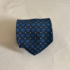Pode incluir: Uma gravata azul com um padrão geométrico repetitivo de quadrados azul-escuros delineados a preto. O design inclui pequenos detalhes quadrados brancos e amarelos posicionados nas interseções da grelha, criando um aspeto estruturado e uniforme.